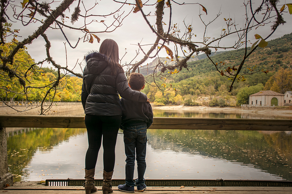 Woman and child stand together on a wooden railing overlooking a lake framed by autumn trees and hills abroad.